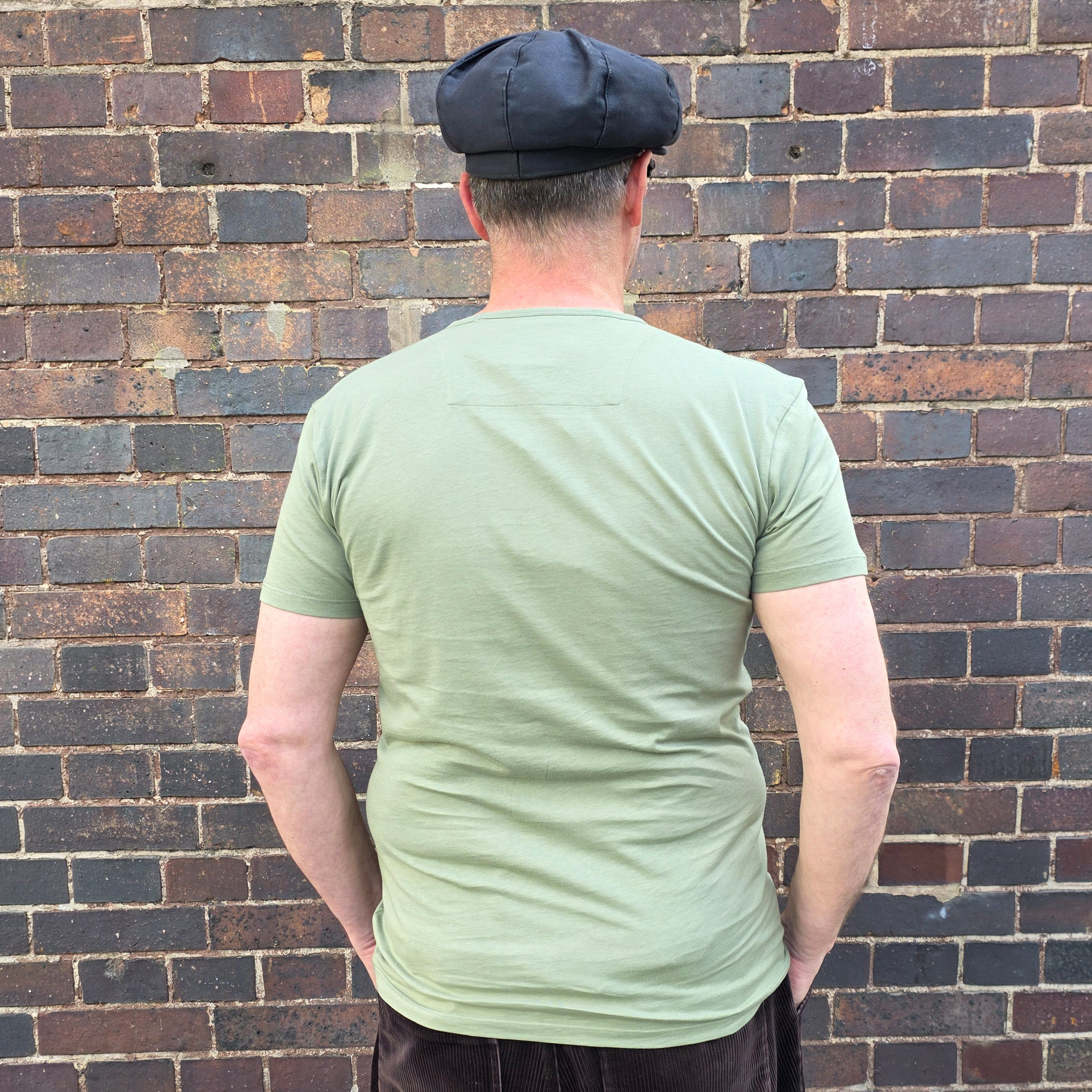 Rückansicht einer Person mit hellgrünem T-Shirt vor einer braun-grauen Backsteinwand.
